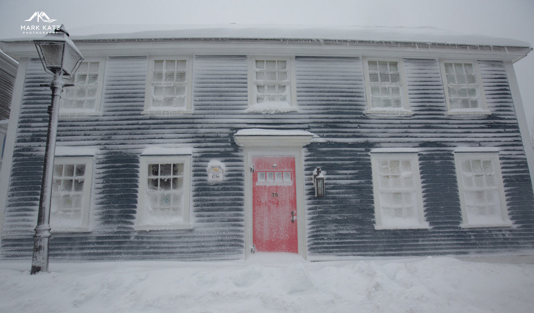 Historic dark clapboard house covered in snow with a red front door and frosted windows in winter light.