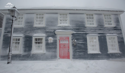 Historic dark clapboard house covered in snow with a red front door and frosted windows in winter light.