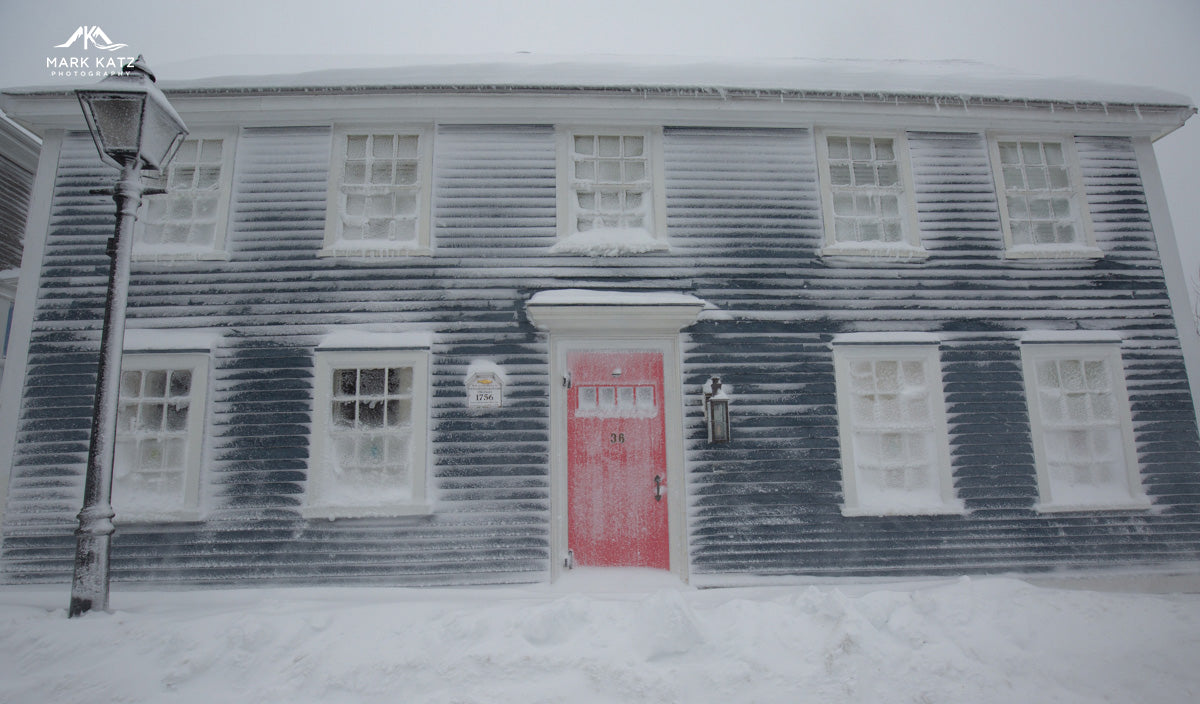 Historic dark clapboard house covered in snow with a red front door and frosted windows in winter light.