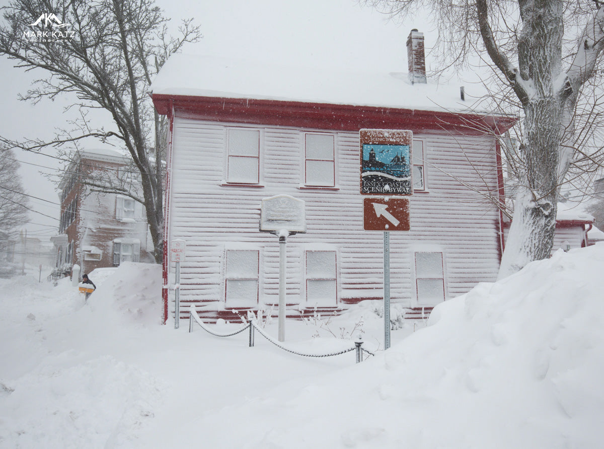 Snow-covered clapboard house with red trim during snowfall in Marblehead, Massachusetts, bordered by snowbanks and signs