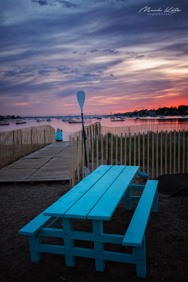A turquoise picnic table and paddle beside a beach path overlooking Marblehead Harbor at sunrise.